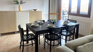 Ein Essbereich mit einem schwarzen Tisch, gedeckt für sechs Personen, umgeben von schwarzen Stühlen. Drei Pendelleuchten hängen über dem Tisch. Ein beiges Sofa ist im Vordergrund teilweise sichtbar.