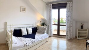Ein Schlafzimmer mit einem weißen Metallbett, einem kleinen Nachttischchen mit Lampe, einer Zimmerpflanze und Blick ins Freie durch ein großes Fenster mit Vorhängen.