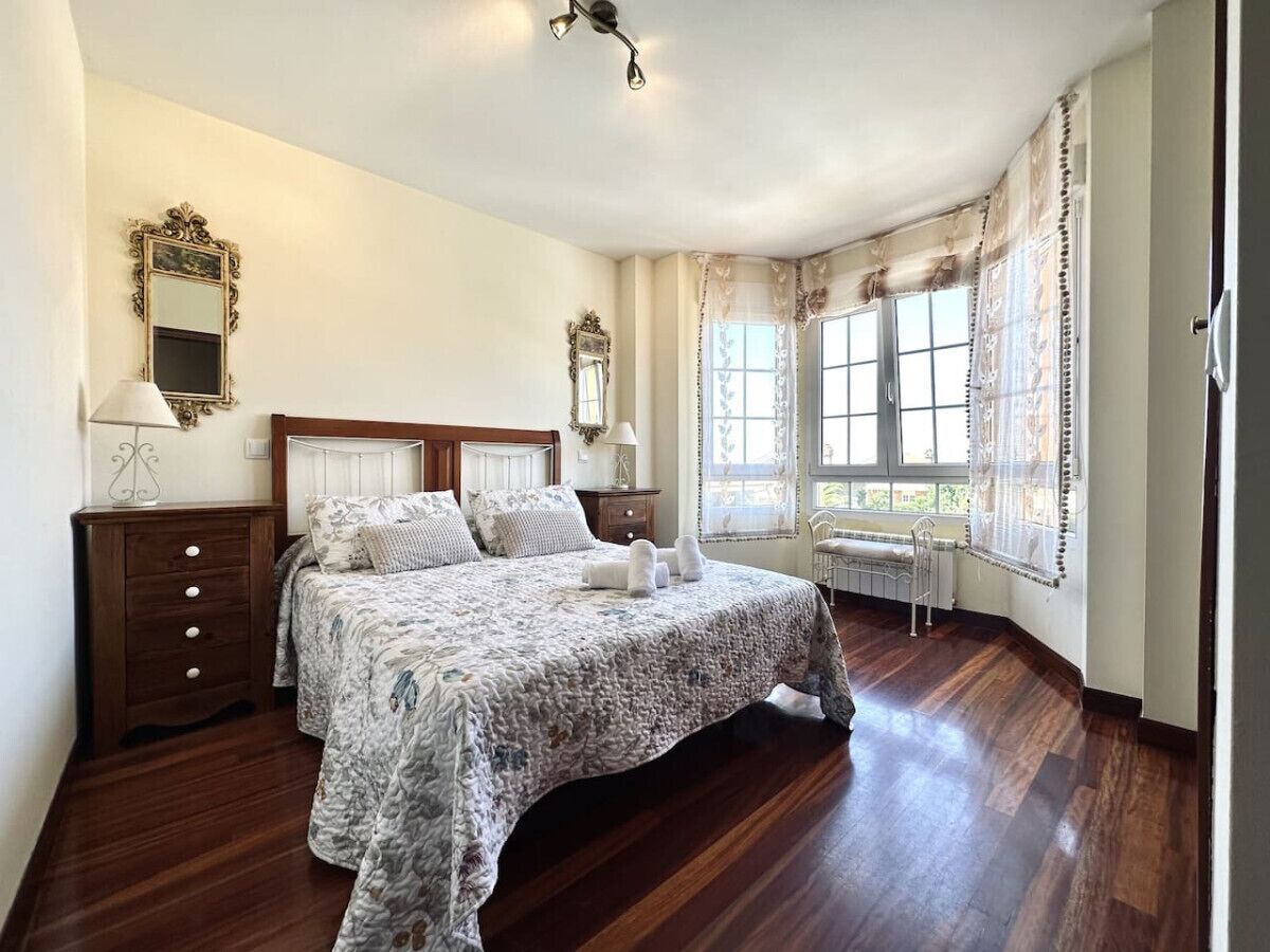 A bedroom with a large bed, wooden floor, two bedside tables, a nightstand, a window with curtains, and a mirror on the wall.
