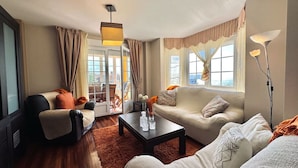 Una sala de estar moderna con un sofá, un sillón y una mesa de centro. Hay un ventanal con cortinas, una lámpara de pie y vistas de la ciudad.