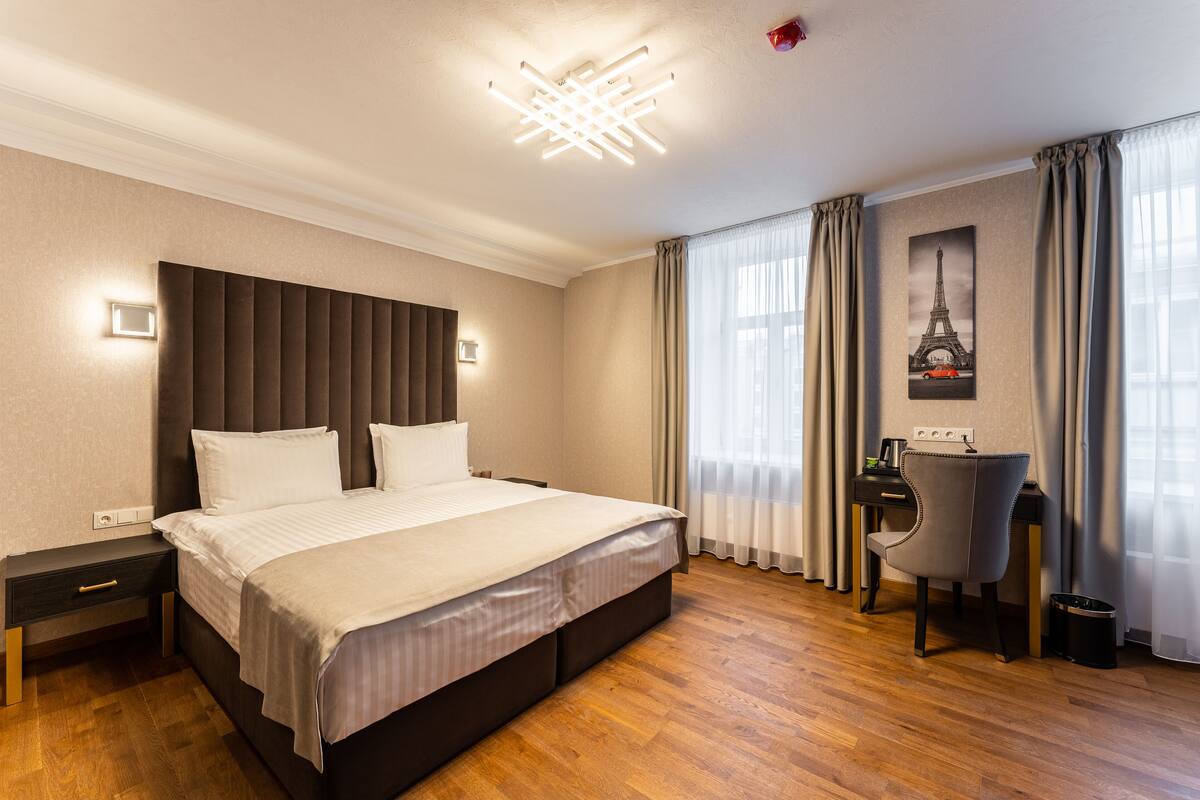 A hotel room with a large bed, a desk, a chair, a picture of the Eiffel Tower, and a modern ceiling light fixture.