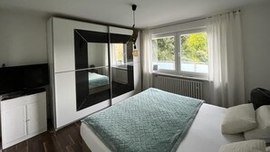 A modern bedroom with a bed, a TV, a wardrobe, and a large window with curtains. - Beautiful vacation apartment in Essen- Burgaltendorf (Essen)