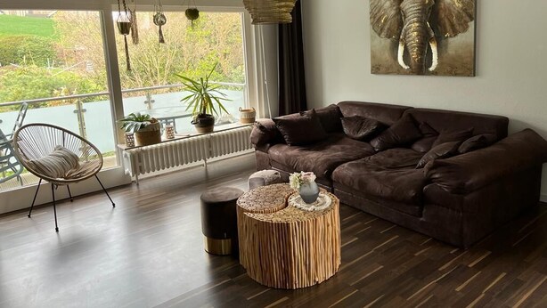 A modern living room with a brown leather sofa, a round woven coffee table, a wicker chair, and a large window with a view of greenery. - Beautiful vacation apartment in Essen- Burgaltendorf (Essen)