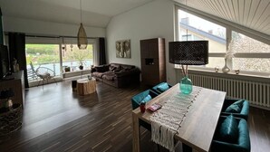 A modern living room with a wooden floor, a dining table with a macrame runner, a sofa, and a chair. There is a large window with a view of greenery outside. - Beautiful vacation apartment in Essen- Burgaltendorf (Essen)