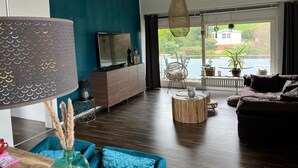 A modern living room with a large TV, a sofa, a coffee table, and a view of a house through large windows. - Beautiful vacation apartment in Essen- Burgaltendorf (Essen)