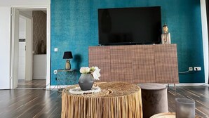 A living room with a teal wall, a flat-screen TV mounted above a wooden cabinet, a round wooden table with a vase of flowers, and a lamp on a side table. - Beautiful vacation apartment in Essen- Burgaltendorf (Essen)