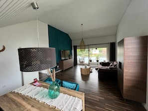 Un salon moderne avec un plancher en bois, une grande lampe noire, un canapé bleu et une vue sur l’extérieur grâce à de grandes fenêtres.