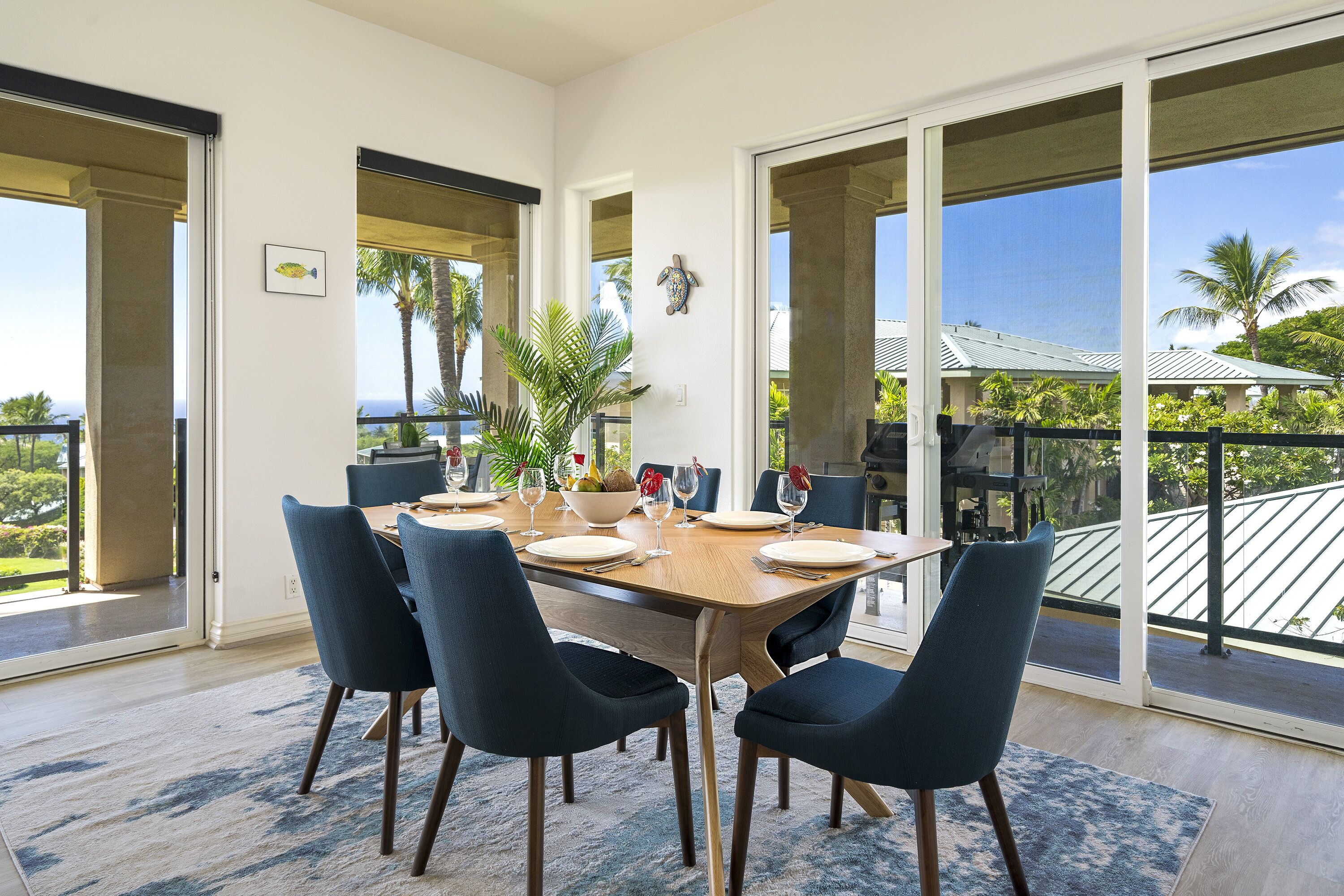 A dining area with a large table set for six, surrounded by dark blue chairs, with a view of the ocean and palm trees through large sliding glass doors.