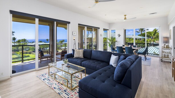A spacious living room with a large sectional sofa, a glass coffee table, and a view of the ocean through large sliding doors.