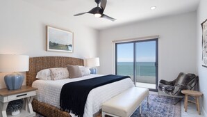 Un dormitorio con una cama grande, un ventilador de techo, vista al mar a través de una puerta corredera y una silla junto a la ventana.
