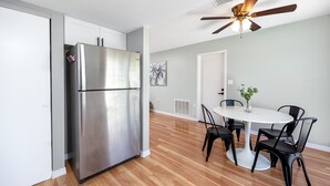 Cozinha moderna com geladeira de aço inoxidável, área de jantar com mesa branca e cadeiras pretas, e um ventilador de teto.