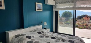 A bedroom with a bed, a nightstand, a lamp, and a window with a view of a garden and mountains.