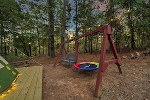 A wooden deck with a swing set and trampoline in a wooded area.