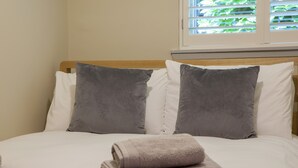 Ein ordentlich bezogenes Bett mit weißen und grauen Kissen, ein gefaltetes Handtuch und ein Fenster mit Jalousien.