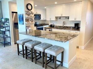 A modern kitchen with a granite countertop, white cabinets, and a stainless steel refrigerator. There are three bar stools and a microwave above the counter.