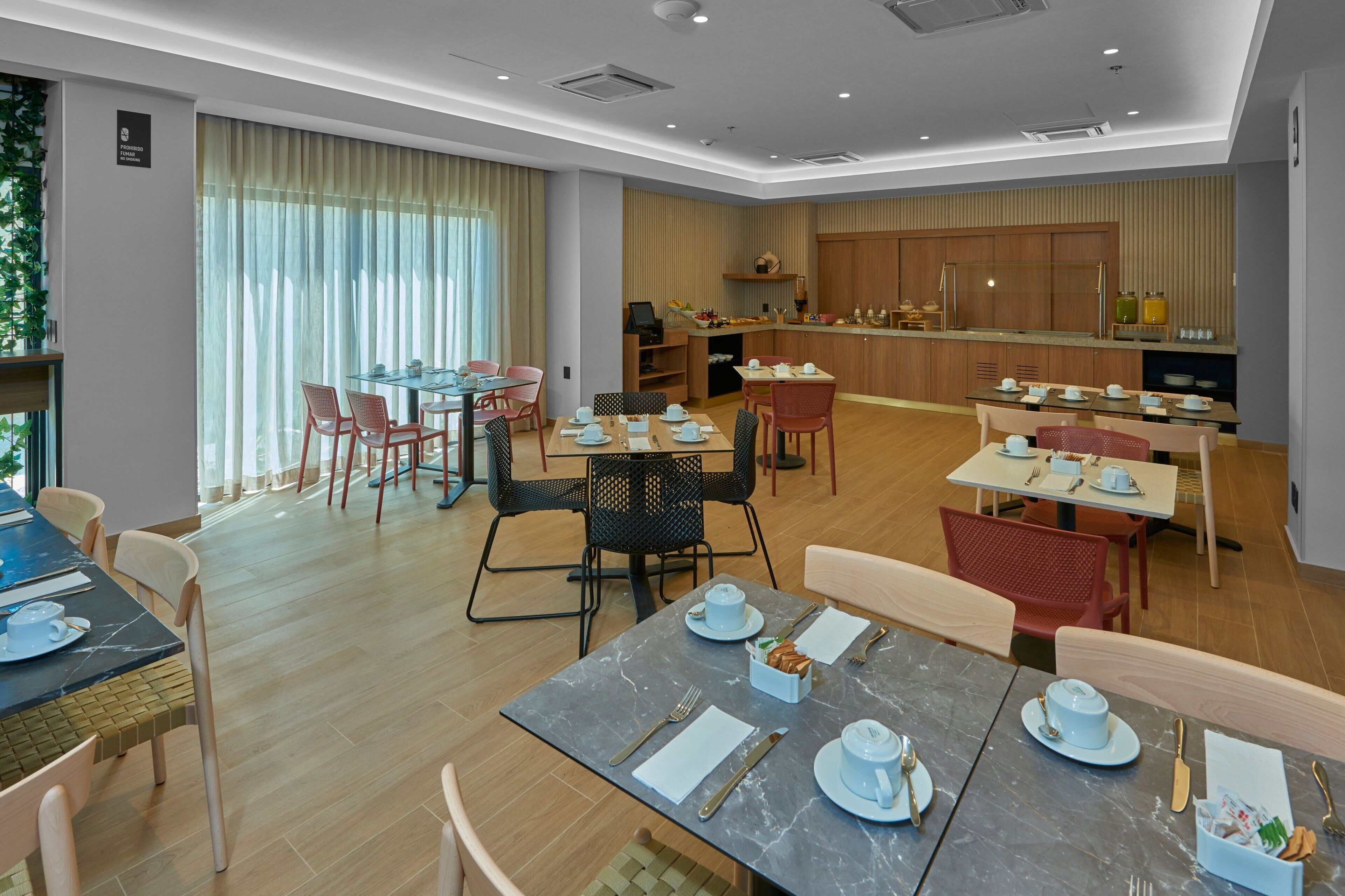 Un espace repas moderne avec plusieurs tables et chaises, une cuisine à l’arrière-plan et une grande fenêtre avec des rideaux.