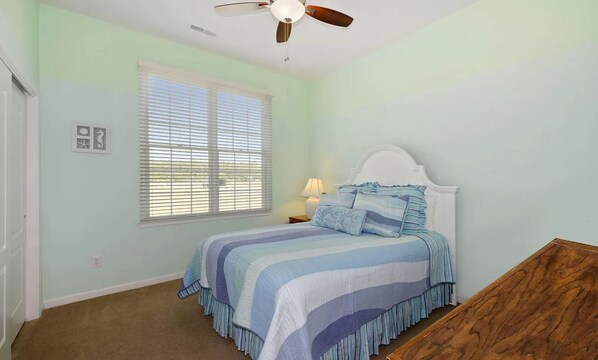 A bedroom with a bed, a ceiling fan, a window with blinds, and a nightstand with a lamp.