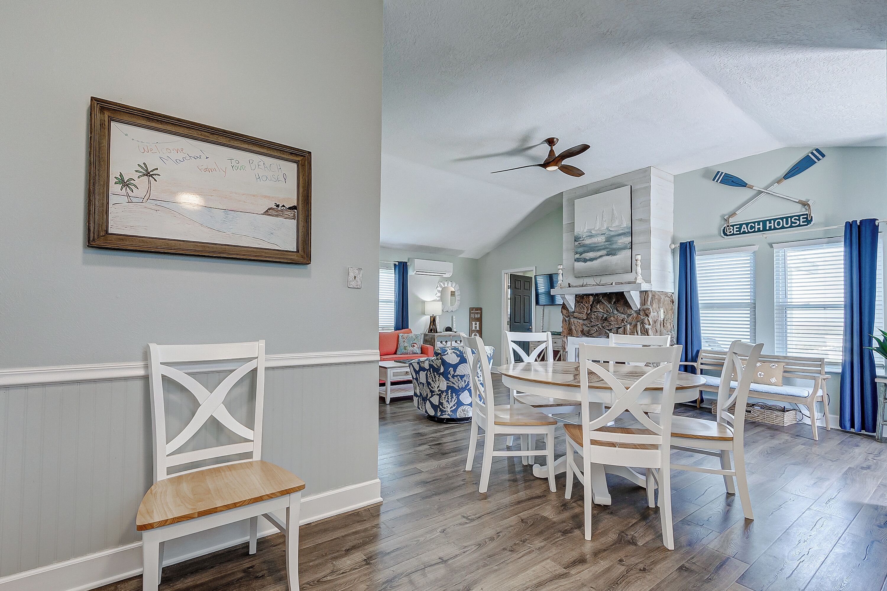 Un coin repas avec des chaises blanches et une table en bois, un ventilateur de plafond et un panneau d’accueil encadré.