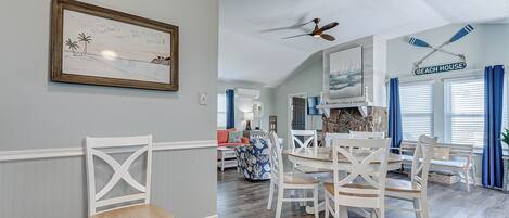 Un coin repas avec des chaises blanches et une table en bois, un ventilateur de plafond et un panneau d’accueil encadré.