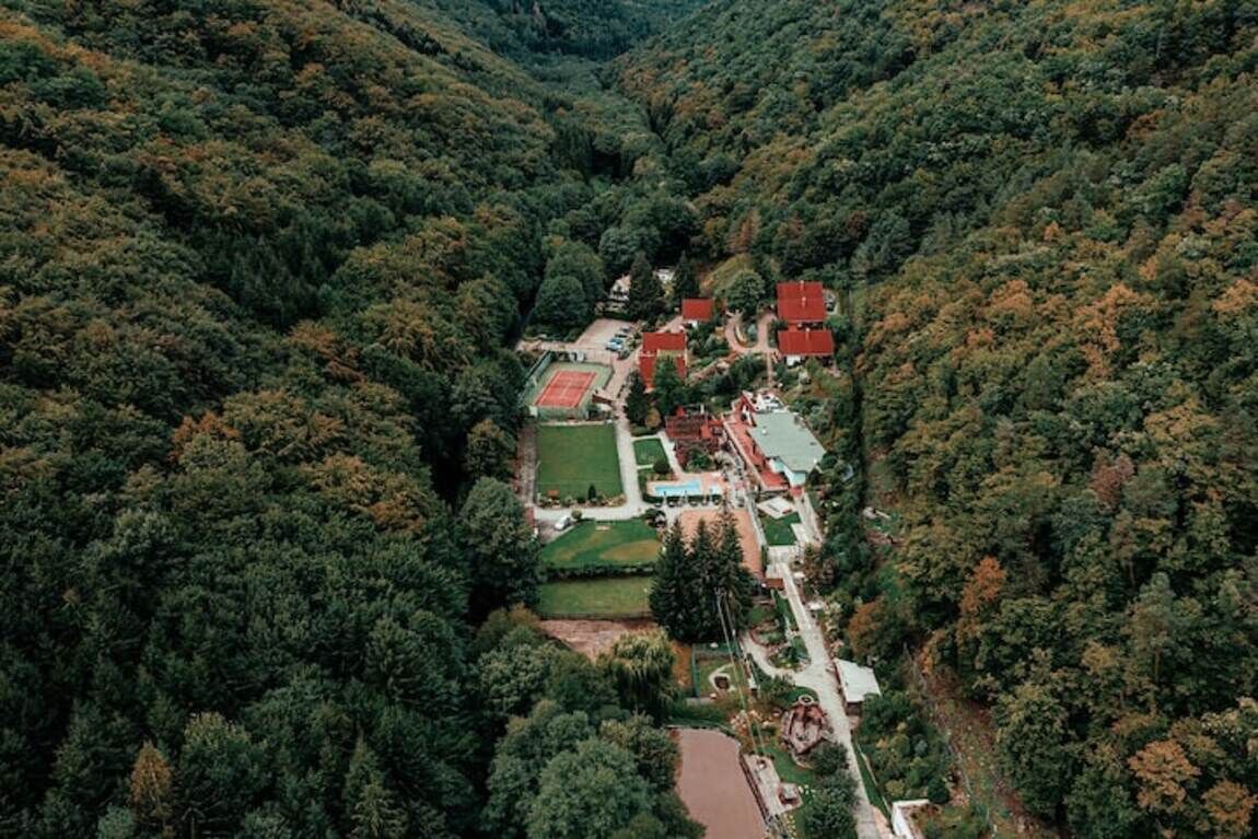 Vista aérea de un pueblo con edificios rodeados de un bosque frondoso, una cancha de tenis y un pequeño río.