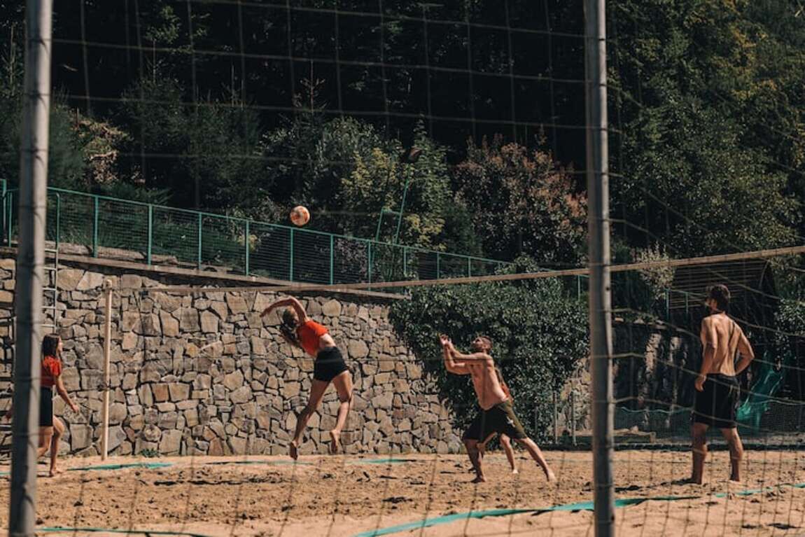 Ein Volleyballspiel auf einem Sandplatz, im Hintergrund eine Steinmauer und Grünpflanzen.