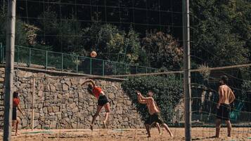 Ein Volleyballspiel auf einem Sandplatz, im Hintergrund eine Steinmauer und Grünpflanzen.