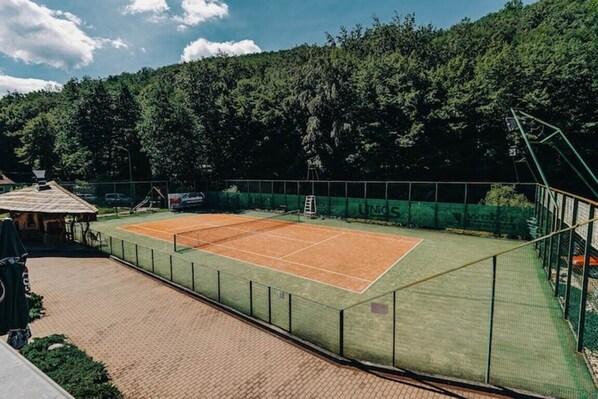A tennis court with a net, surrounded by a green fence, and a brick pathway leading to a building.