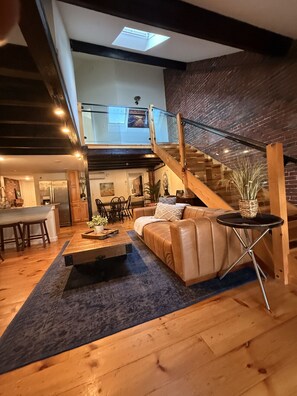 Amplia sala de estar con una mesa de centro de madera, un sofá de cuero y una pequeña mesa redonda con una cesta tejida. Una escalera con barandilla de cristal conduce a un nivel superior.