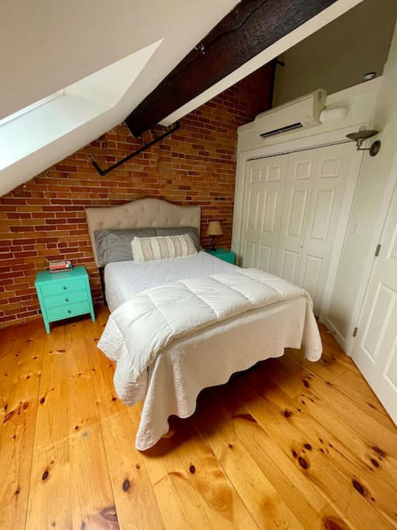 A bedroom with a sloped ceiling, exposed brick walls, a wooden floor, a bed with a white comforter, a nightstand, and a door.