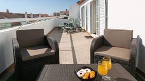 A balcony with wicker furniture, a table with a fruit bowl and drinks, and a view of buildings in the background. - Peniche Terrace - Spacious apartment in the center (Peniche)
