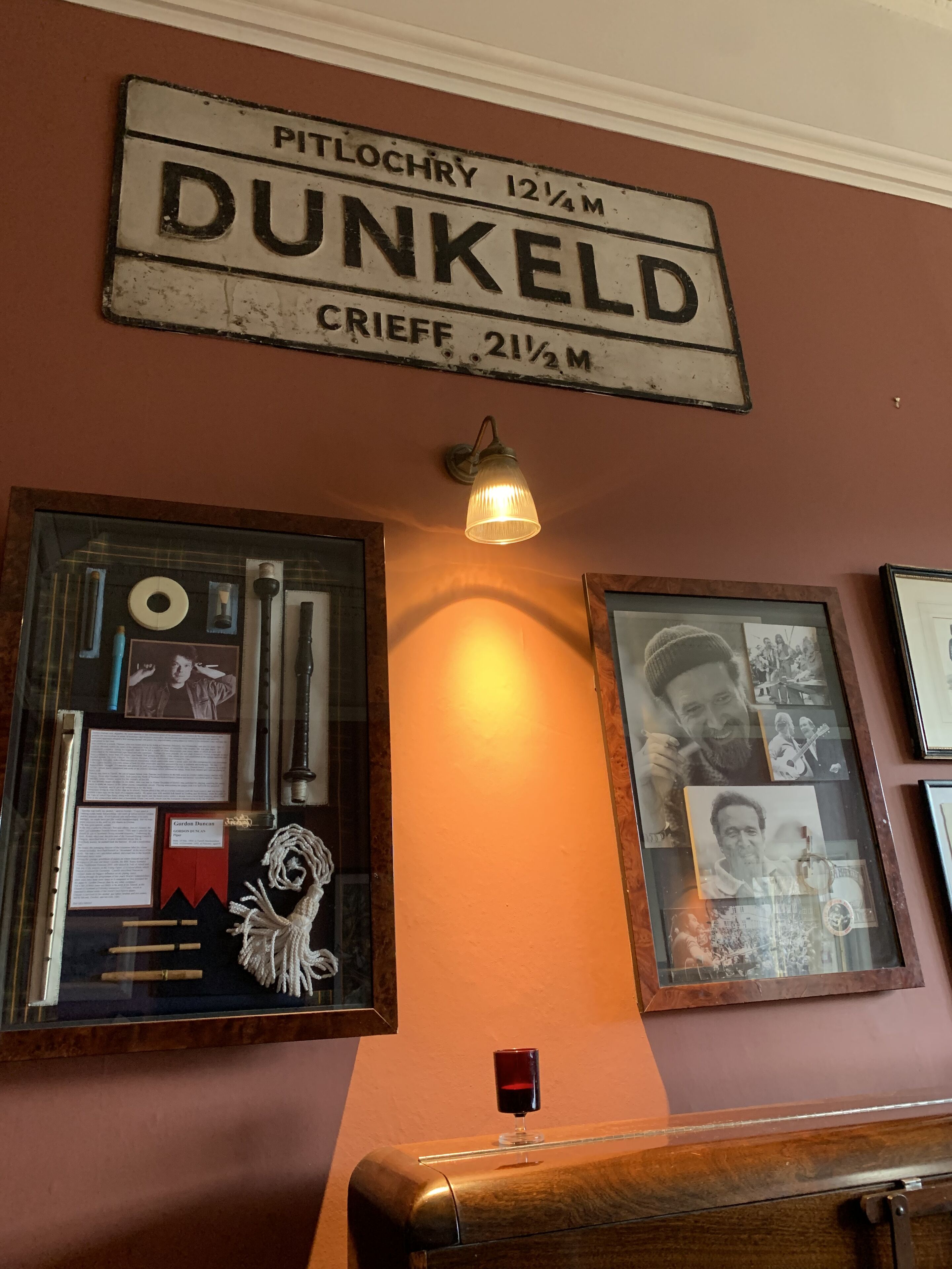 A wall with a sign reading "DUNKELD" and a framed display of various items including photographs and documents.