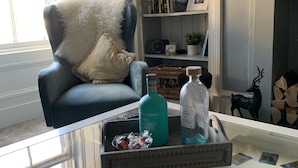 Una sala de estar con un sillón azul, una mesa de centro de vidrio con revistas, una estantería y una chimenea.