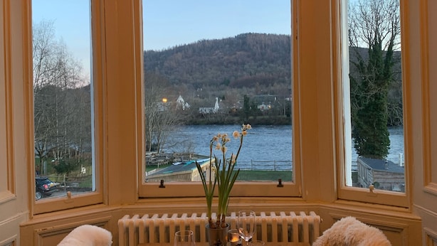 Una mesa de comedor con sillas, un jarrón con flores y vistas a un lago y bosque a través de amplios ventanales.
