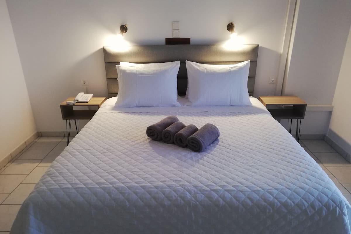 Una cama bien hecha con un edredón blanco acolchado, dos almohadas blancas y toallas enrolladas sobre la cama. Hay dos mesitas de noche con lámparas, un teléfono y un pequeño atril de madera.
