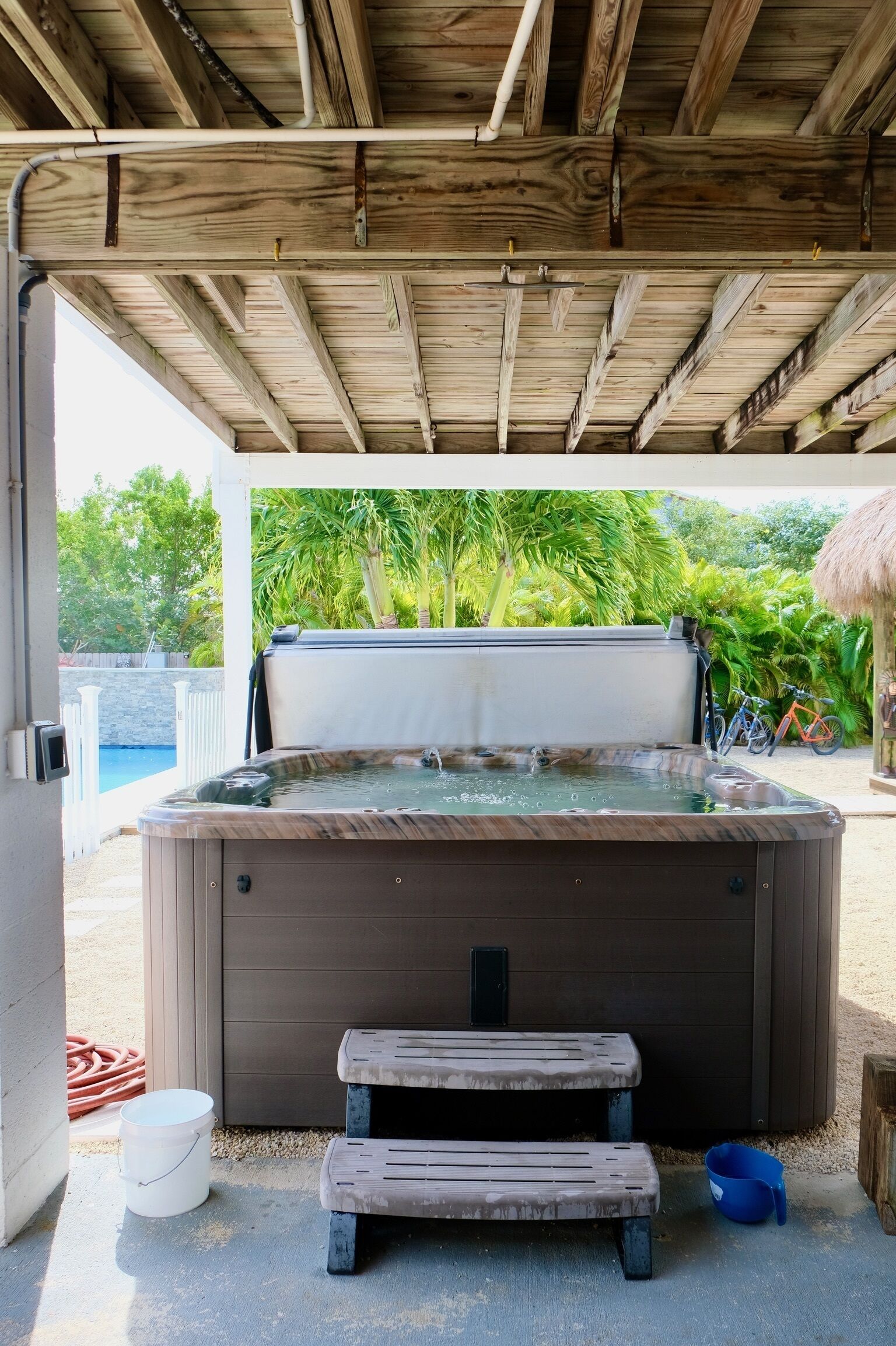 A hot tub under a wooden pergola with a view of a swimming pool and greenery.