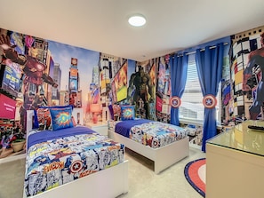 Een slaapkamer met twee bedden, elk voorzien van een dekbedovertrek met stripboekthema. De kamer heeft een grote muurschildering van een stadsgezicht met superhelden op de muren.