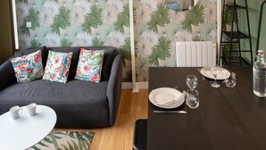 A modern living room with a grey sofa, floral cushions, a small white table, and a dining area with a dark table set for two. - Canopée - Lovely studio with A/C Rhône riverbank (Lyon)