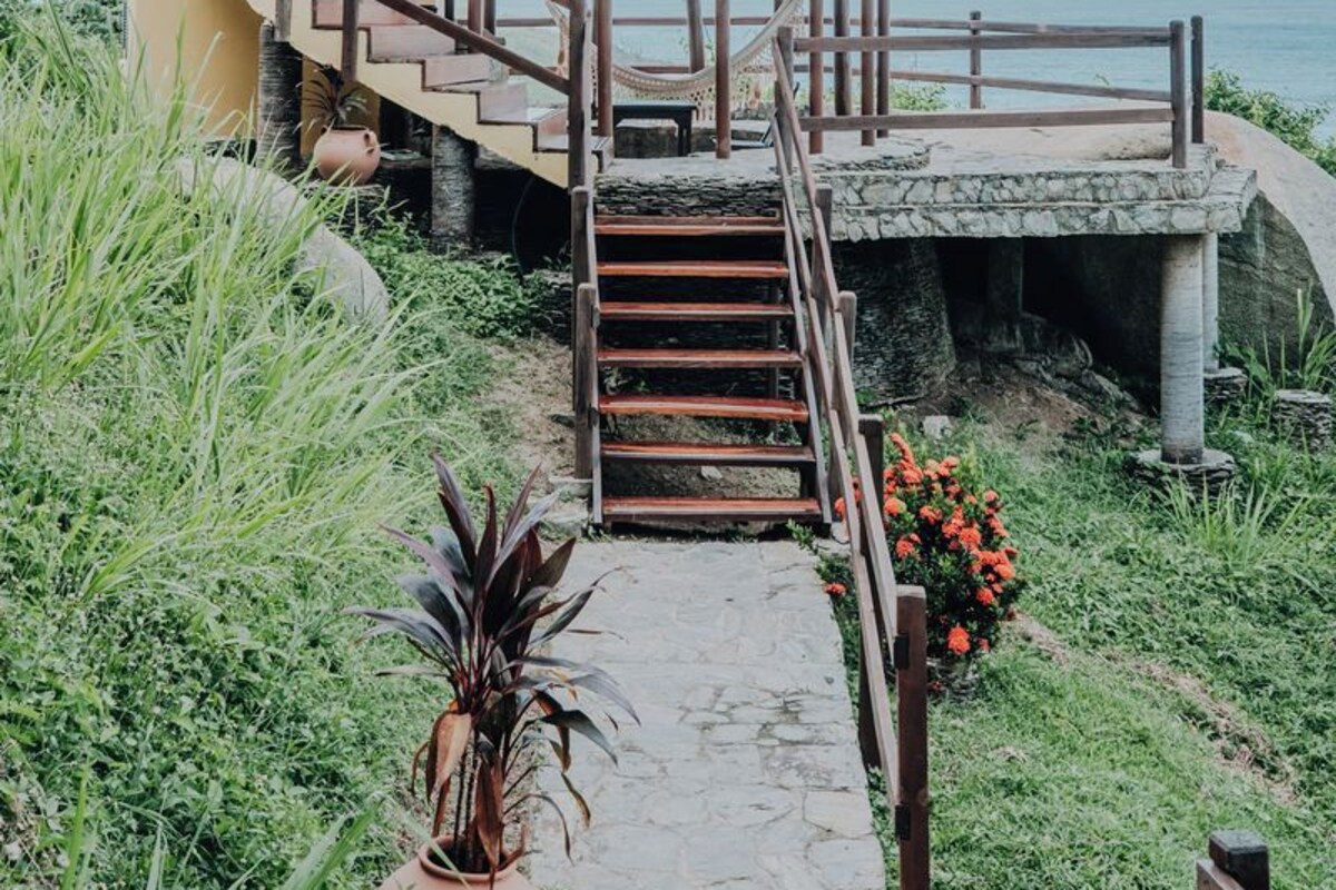 Una estructura con techo de paja y una escalera de madera que conduce a una plataforma, rodeada de vegetación y con vista a un cuerpo de agua.
