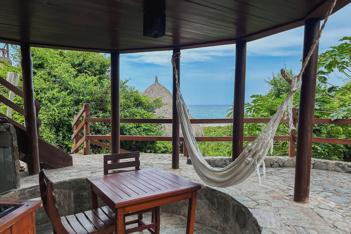 A wooden table and chairs under a wooden roof, a hammock, and a view of the ocean.