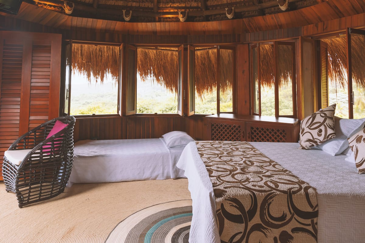 A room with a bed, a wicker chair, and a view through large windows.