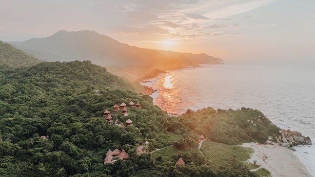 An aerial view of a coastal area with lush greenery, a beach, and several thatched-roof huts.