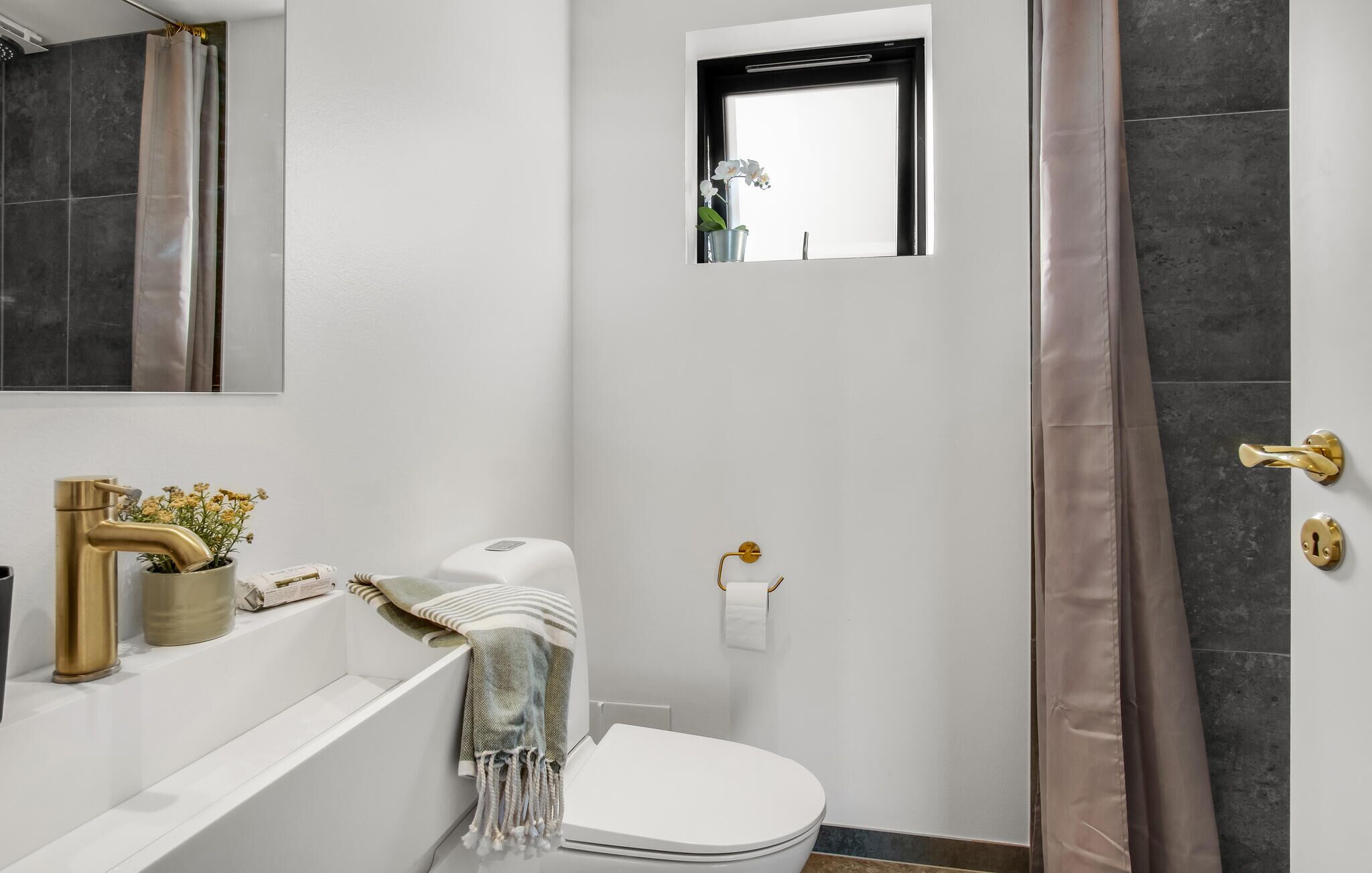 Ein Badezimmer mit Toilette, Handtuchhalter, kleinem Fenster, Duschvorhang und Waschbecken mit goldfarbener Armatur.