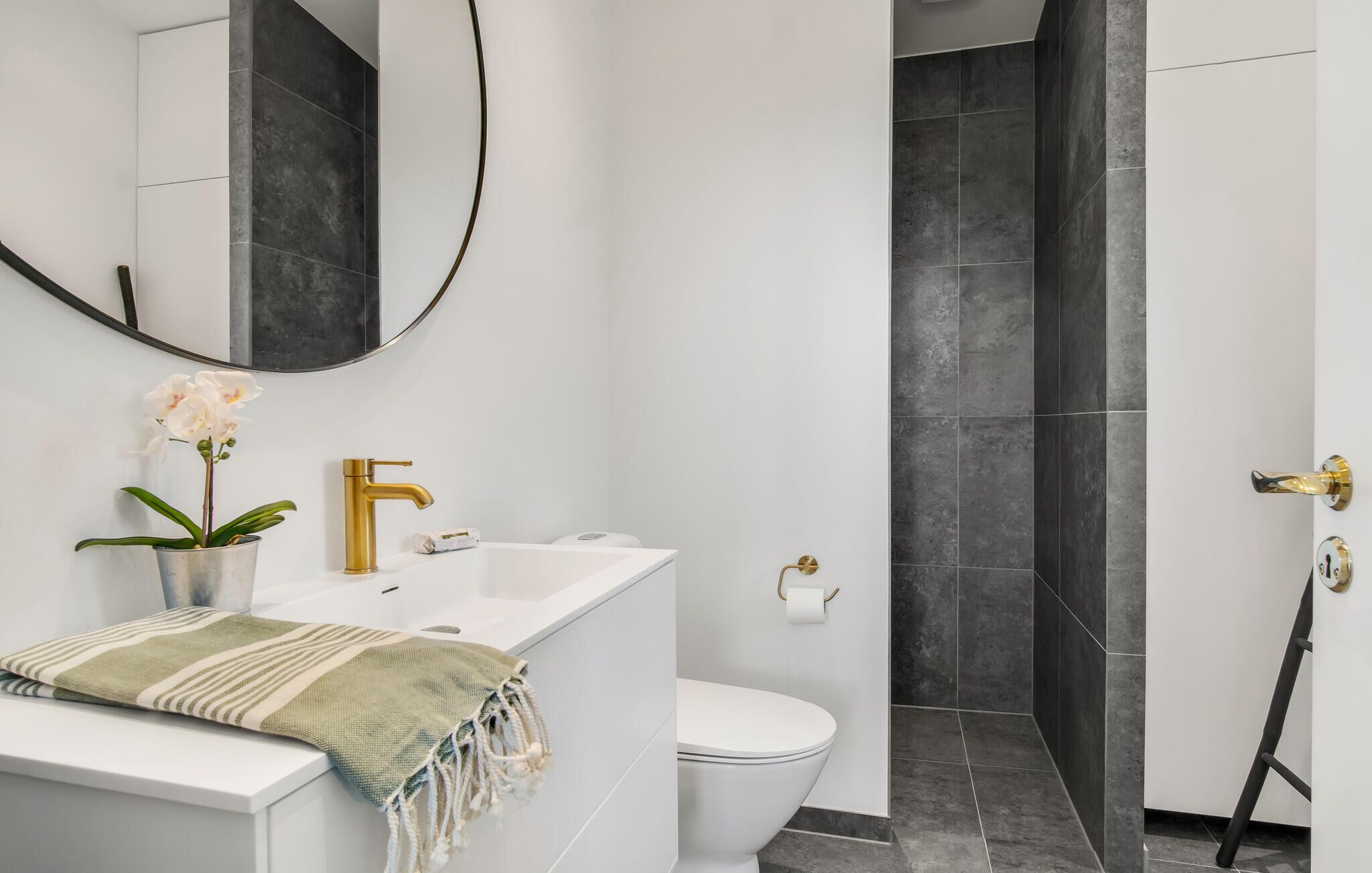 Een moderne badkamer met een witte wastafel, gouden details, een potorkidee en een douche met donkere tegels.