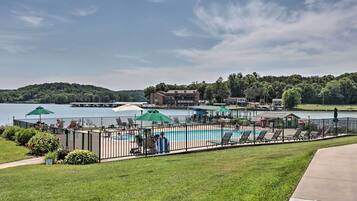 A pool area with lounge chairs and umbrellas, surrounded by a fence, with a lake and buildings in the background.