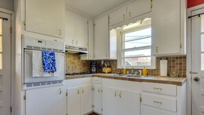 Une cuisine avec des meubles blancs, un four encastré, une fenêtre et un pare-feu en carreaux.