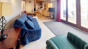 Un salon avec un canapé, une table et un téléviseur. On aperçoit un coin cuisine au fond.