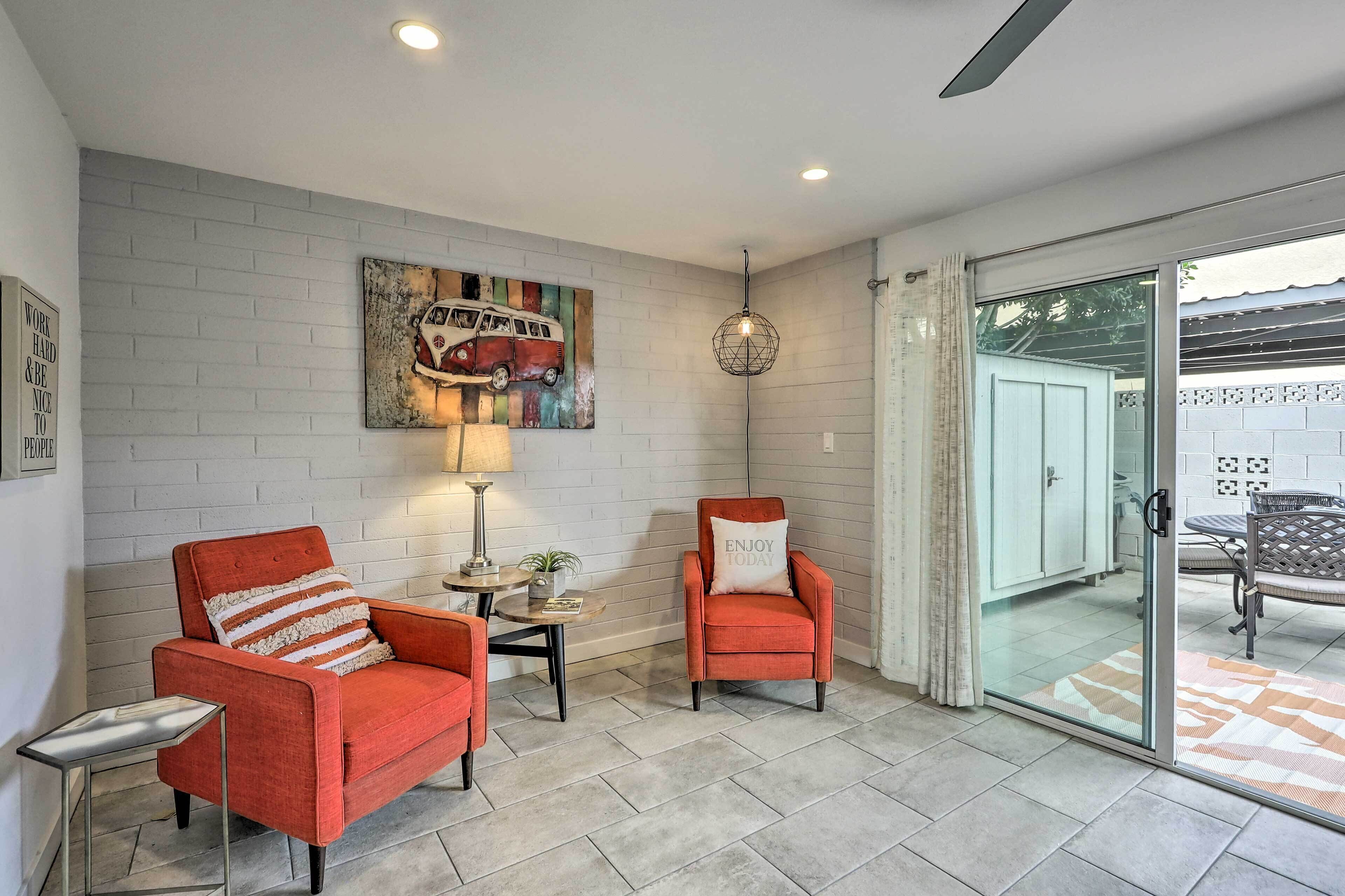 A modern living room with two red armchairs, a coffee table, and a wall-mounted lamp. A large painting of a vintage car decorates the wall, and a sliding glass door leads to an outdoor area.