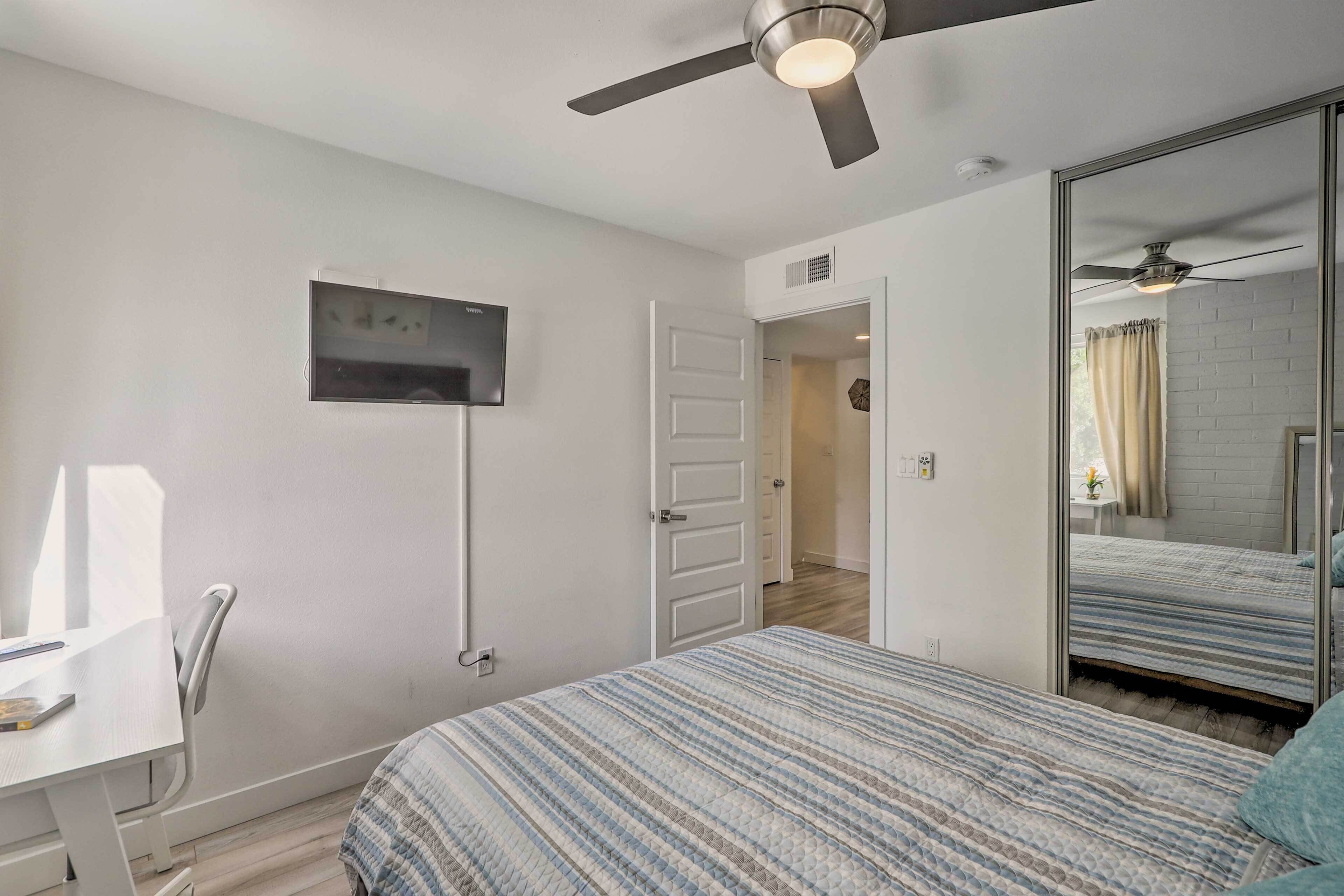 A bedroom with a bed, a desk, a ceiling fan, a TV, and a mirror.
