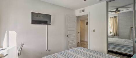 A bedroom with a bed, a desk, a ceiling fan, a TV, and a mirror.