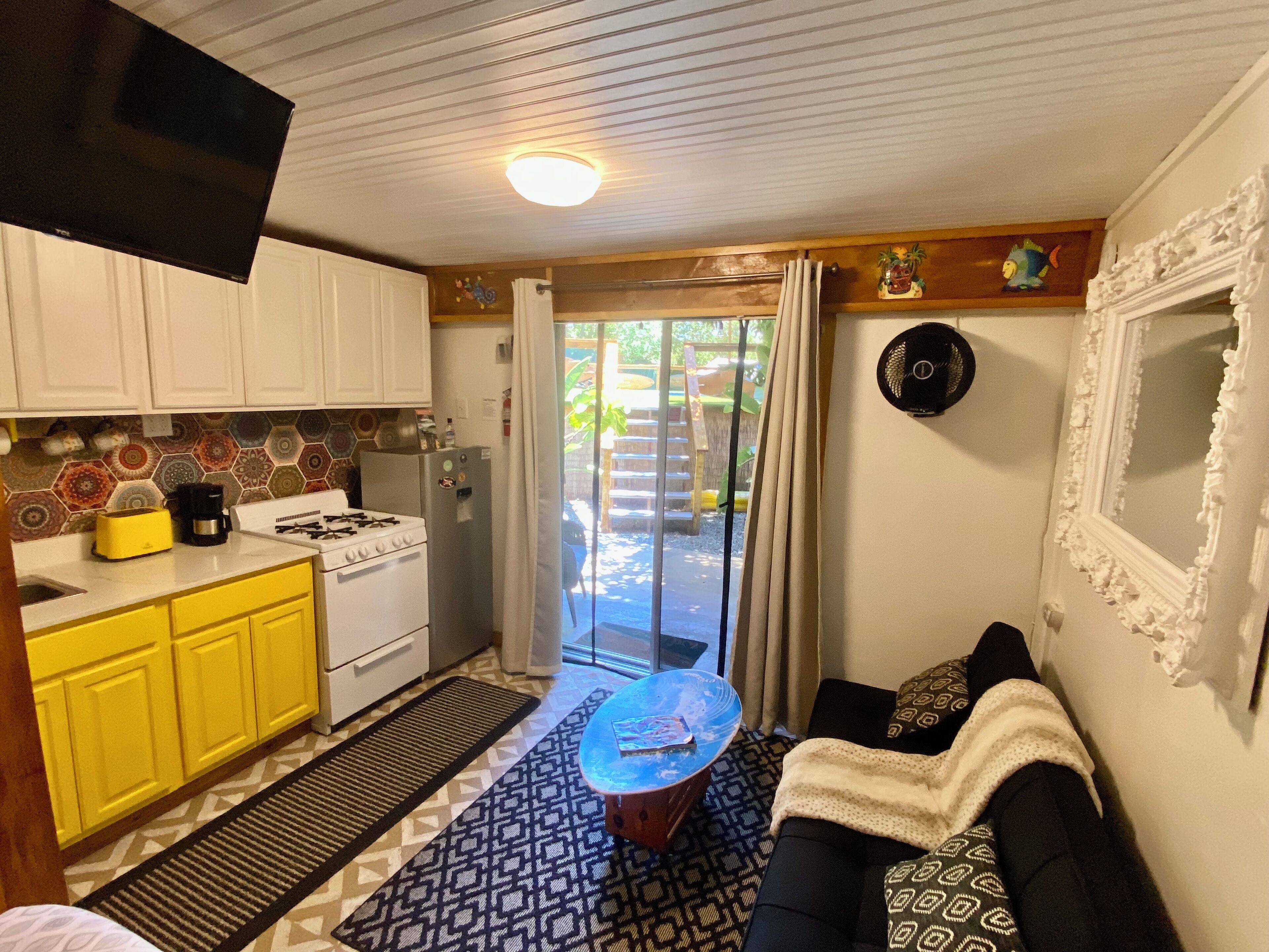 A compact kitchen with white cabinets, a yellow toaster, and a stove. There's a patterned rug and a black sofa with patterned cushions. A small table with a glass top is in the center.